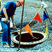 Worker with red helmet looking down a manhole, with safety flags in foreground. By National Institute for Occupational Safety and Health.